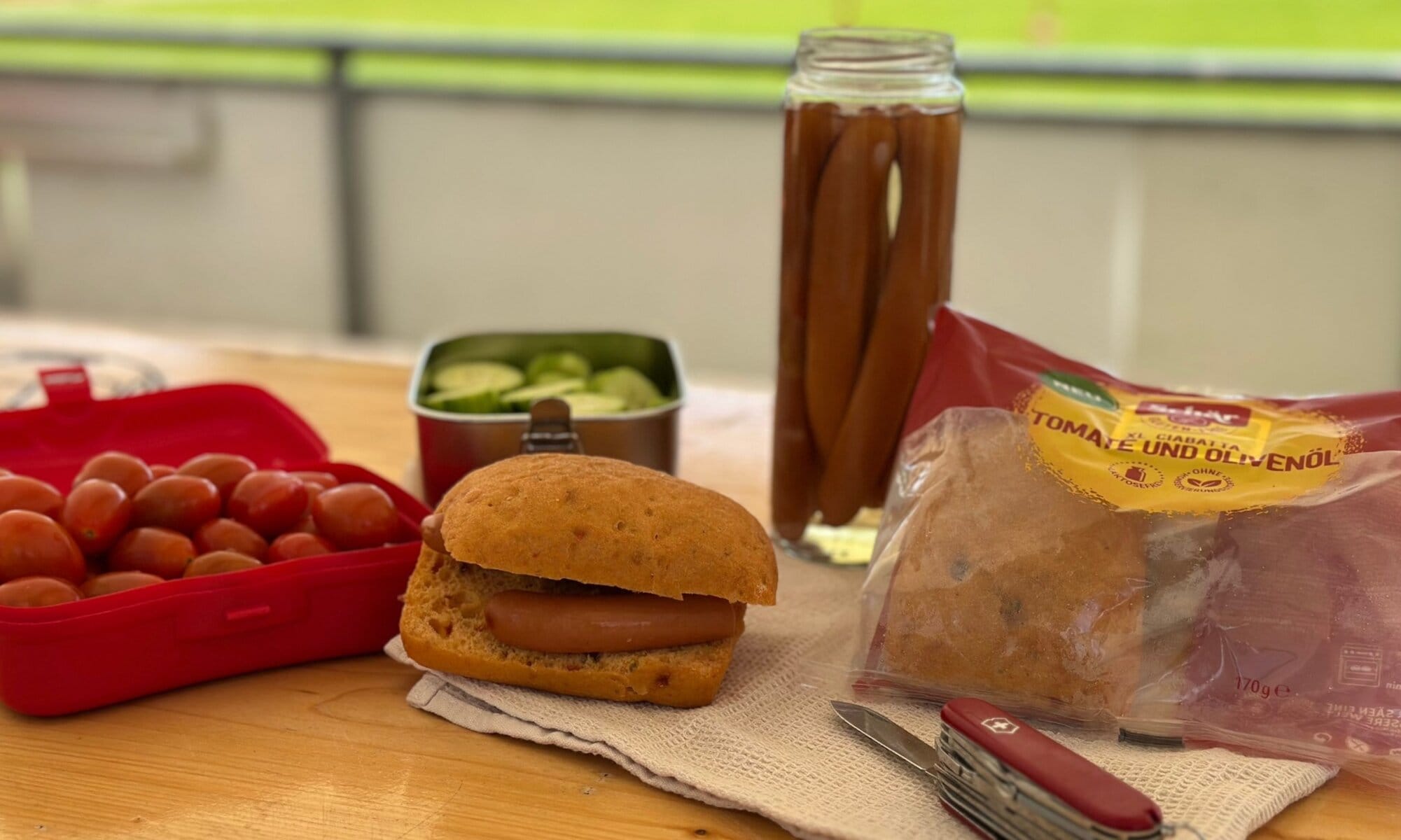 Glutenfreies Grillbrötchen mit Würstchen, Gemüse & Schär-Ciabatta Angerichtetes glutenfreies Brötchen mit Würstchen, Gurkenscheiben, Cocktailtomaten und Schär Ciabatta-Packung im Hintergrund