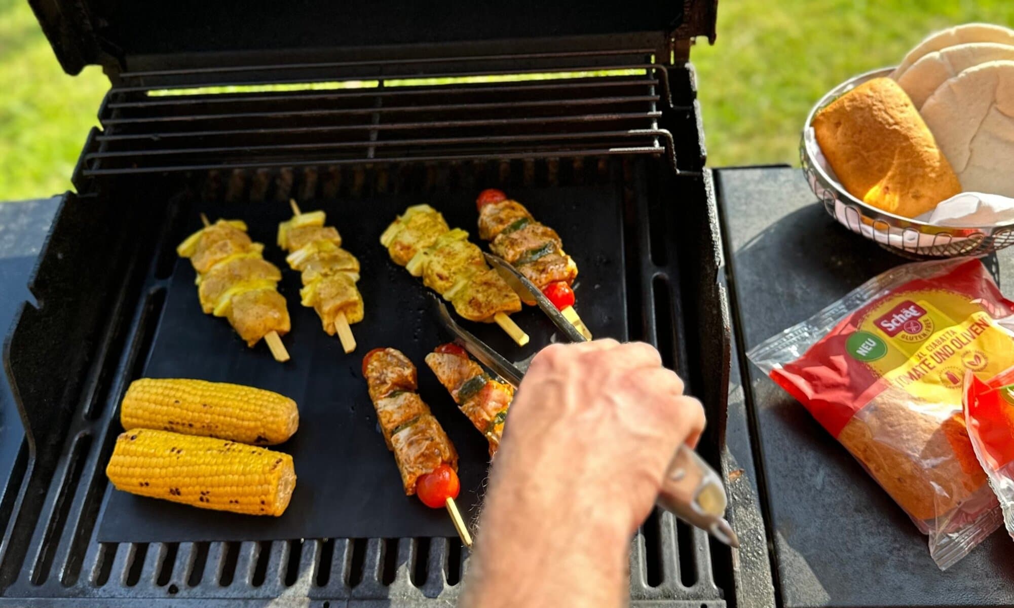 Glutenfrei grillen mit Grillmatte und Schär-Brötchenauswahl Grill mit Grillmatte, Maiskolben und Spießen – daneben Brotkorb mit glutenfreien Ciabatta-Brötchen und Pitas von Schär