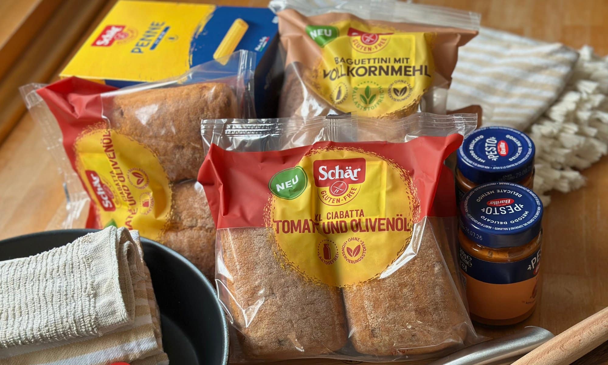 Glutenfreier Proviant und Küchenausstattung für den Selbstversorgerurlaub mit Kindern: Schär-Brötchen, Schär-Nudeln, Pesto, Pfanne, Geschirrlappen, Tischdecke, Kochlöffel, scharfes Messer und Schneidebrett.