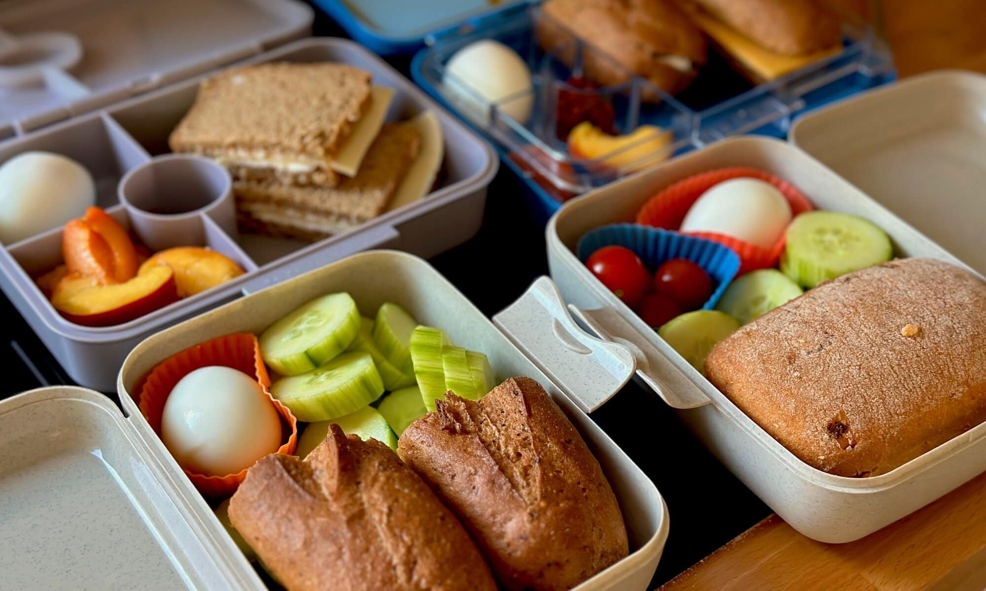 Geöffnete Brotdosen mit glutenfreien, geschmierten Brötchen, hartgekochten Eiern, Cocktailtomaten, Gurkensticks und Pfirsichstücken – vorbereitet für den glutenfreien Selbstversorgerurlaub mit Kindern.