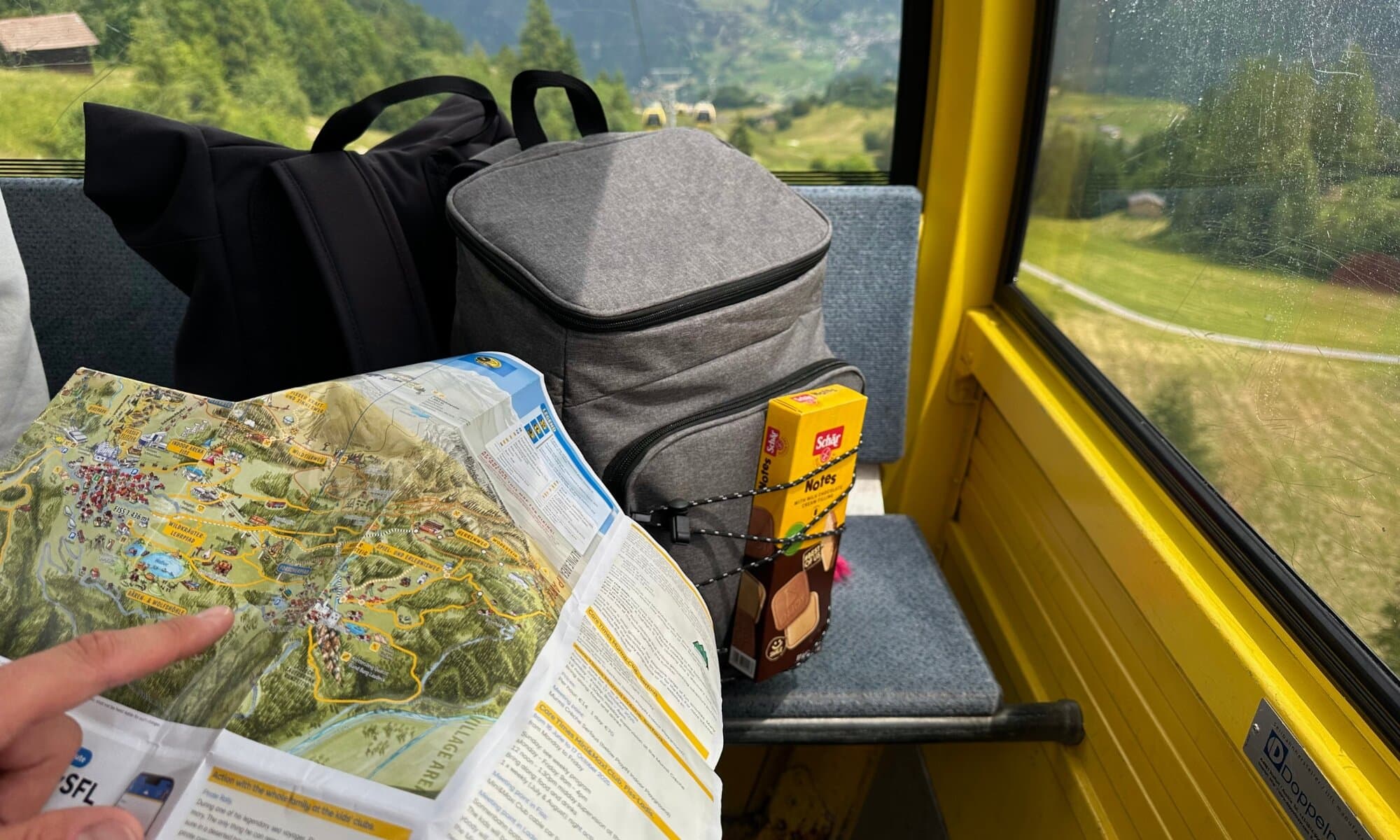 Blick aus einer Gondel auf die Berglandschaft. Im Vordergrund ein Rucksack mit glutenfreien Schär-Produkten und einer Wanderkarte für den nächsten Ausflug während des glutenfreien Selbstversorgerurlaubs mit Kindern.