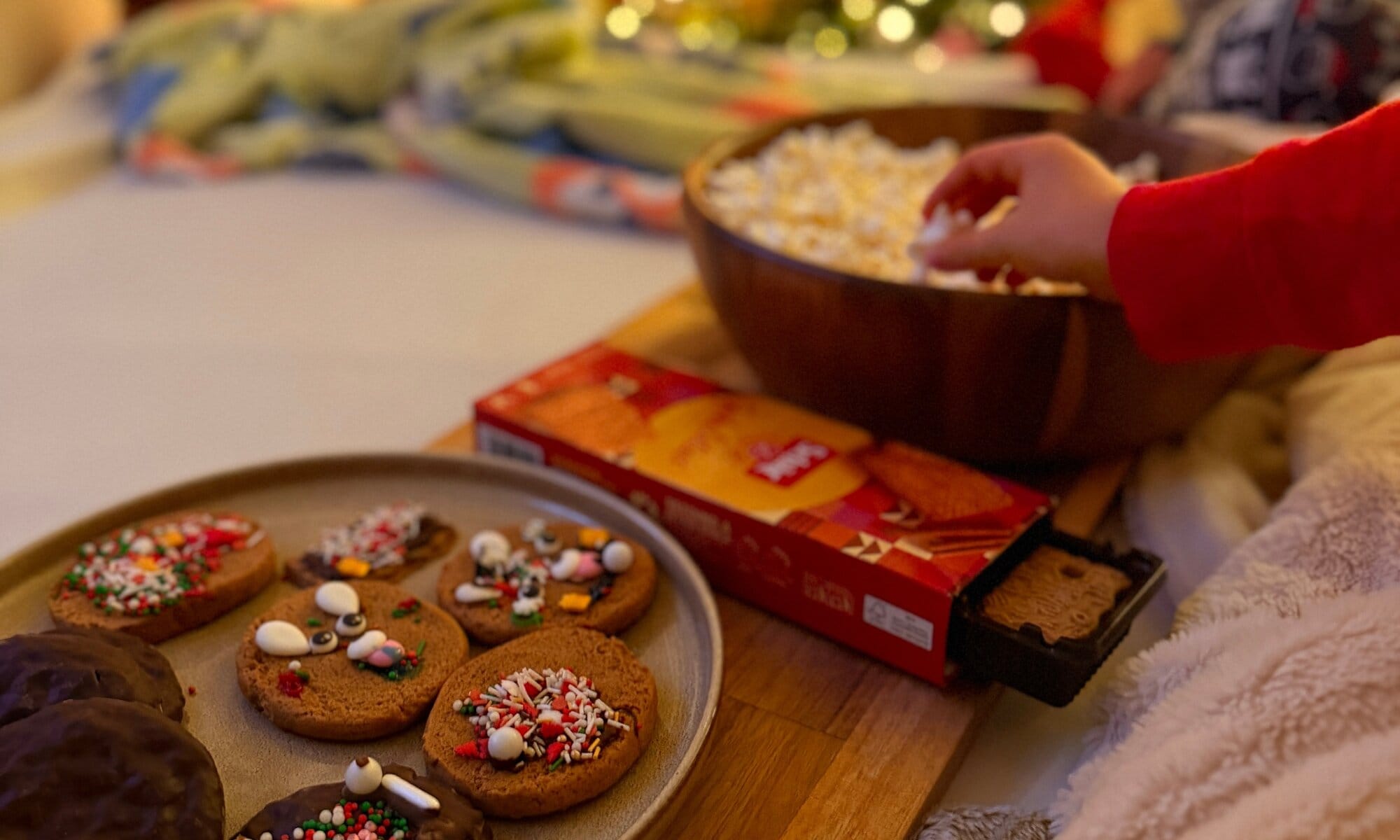 Holzschüssel mit Popcorn, in die eine Kinderhand greift, daneben eine geöffnete Packung glutenfreier Schär-Spekulatius sowie selbst dekorierte glutenfreie Lebkuchen und Cinnamon Soft Cookies.