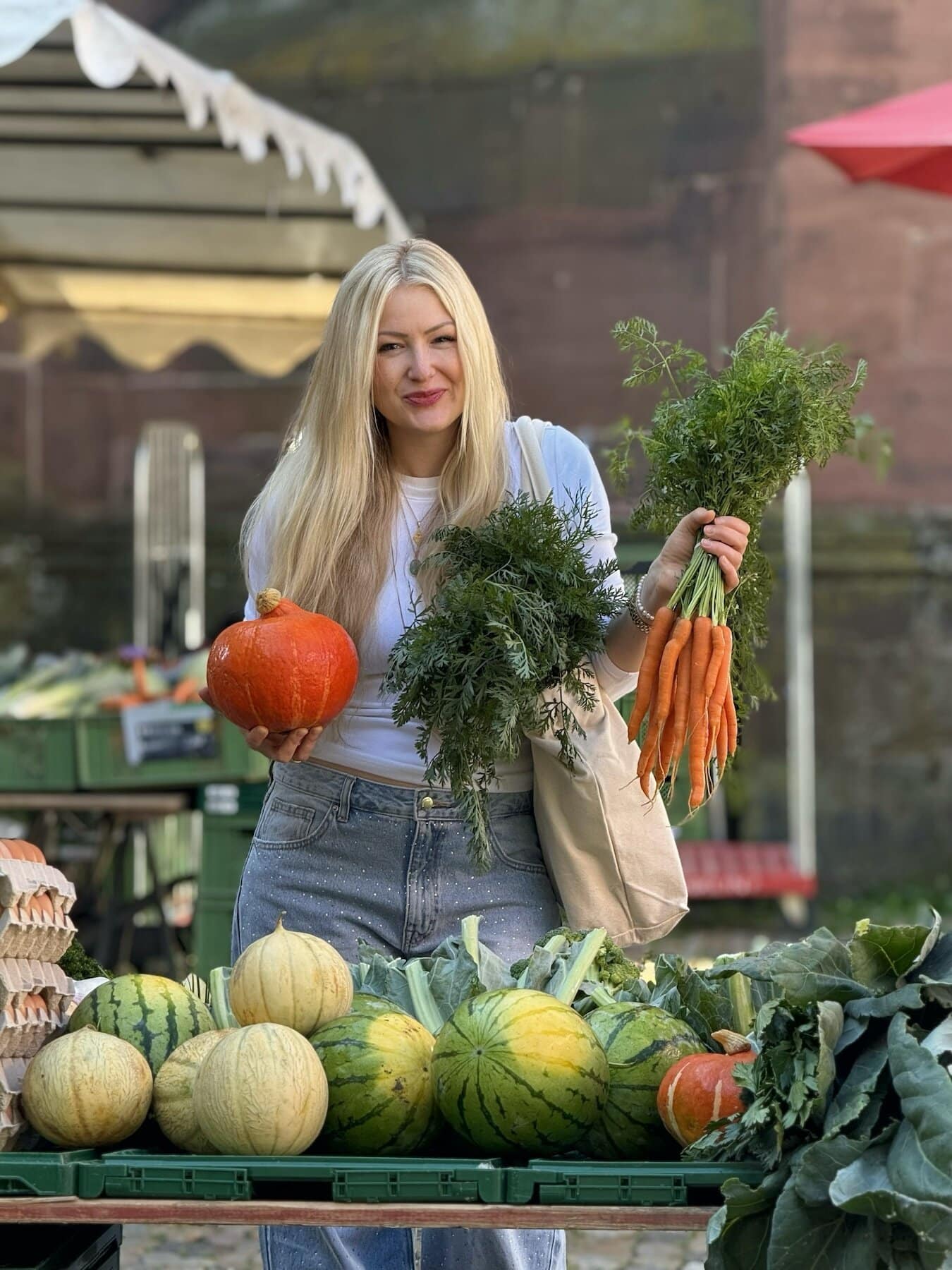 Aurélie steht auf dem Wochenmarkt am Freiburger Münster und hält frisches Gemüse wie Karotten und Kürbis in den Händen